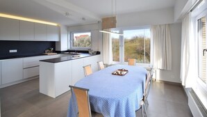 Une cuisine moderne dotée d’un îlot central, d’un coin repas avec une table et des chaises, et de grandes fenêtres offrant une vue sur l’extérieur.