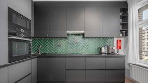 A modern kitchen with dark cabinetry, a green herringbone backsplash, and a window with a view of buildings. - Renovated Apartment for Family (Blankenberge)