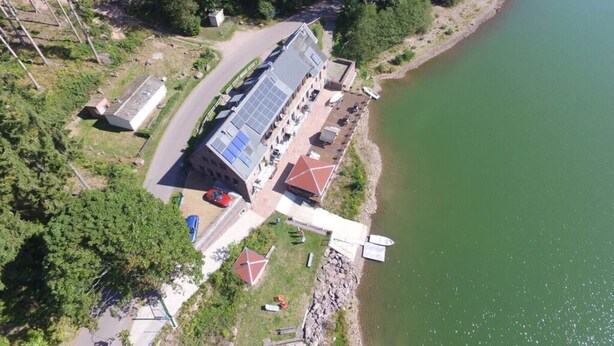 An aerial view of a building with a long, narrow structure, a covered outdoor area with seating, and a dock extending into a green body of water. - Kickelhahn Comfortable Holiday Residence (Geratal)