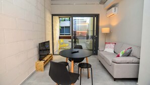 A compact living space with a sofa, a small round table, two black chairs, a television on a wooden stand, and a sliding glass door leading to an outdoor area with a yellow chair.