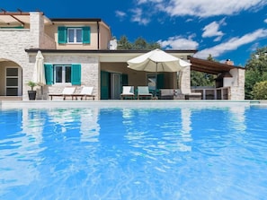 A poolside area with a clear blue pool, a stone building with green shutters, and a pergola with a white umbrella. - Villa Martha Comfortable Holiday Residence (Zminj)