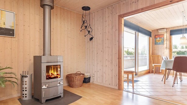 A cozy room with a wood-burning stove, wooden walls, a dining area with chairs and a table, and a view through a glass door.