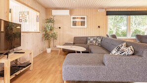 A modern living room with a grey sectional sofa, a flat-screen TV, a wooden TV stand, a potted plant, and a large window offering a view of trees.
