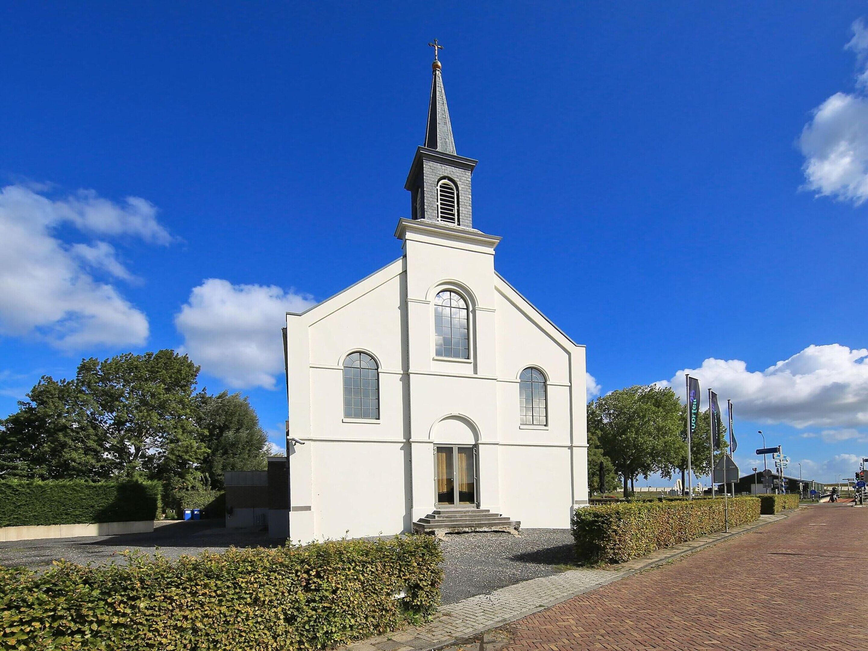 A small white church with a steeple and arched windows, surrounded by hedges and a brick pathway.