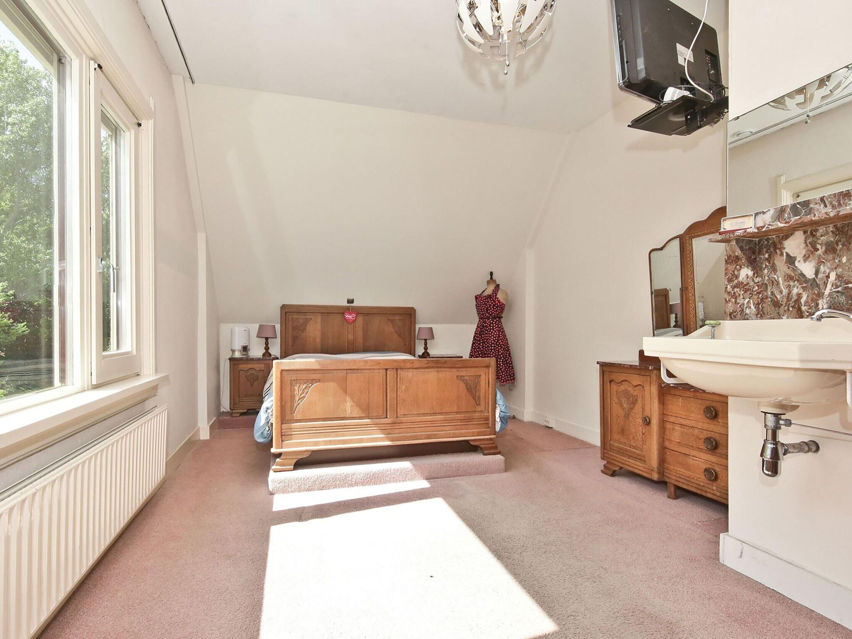 A bedroom with a wooden bed, a dresser, a vanity with a mirror, and a window with a view of trees.