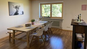 A dining area with a wooden table, white chairs, a window with a view of greenery, and a radiator below. - Appealing Apartment Near Lake (Argenbühl)