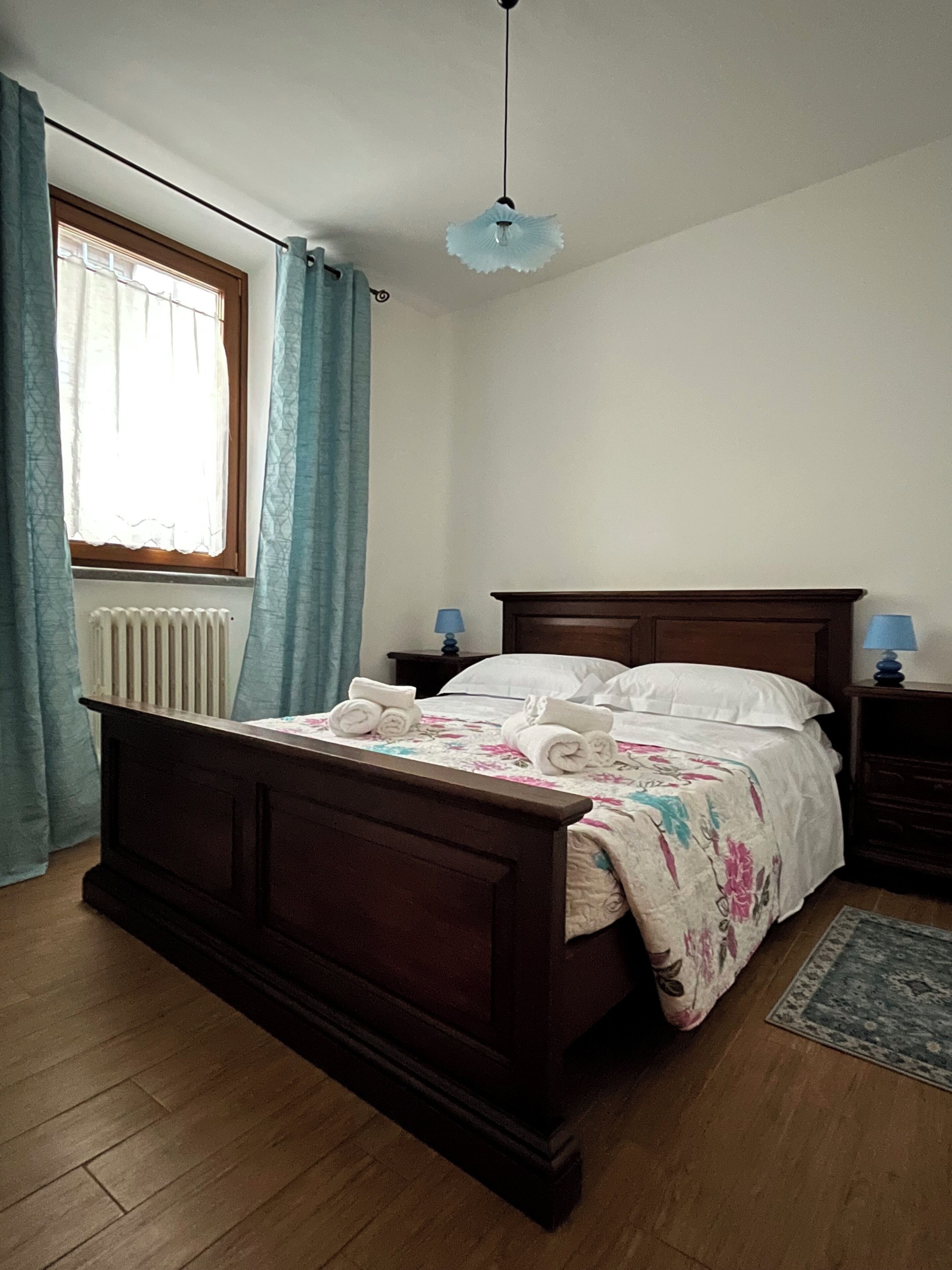 A bedroom with a wooden bed, a nightstand, a lamp, a radiator, and a window with curtains.