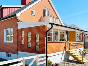 Ein rotes Backsteinhaus mit weißer Fassade, einer Holztreppe, die zu einer Veranda führt, und einem kleinen Außenbereich mit Sitzgelegenheit.