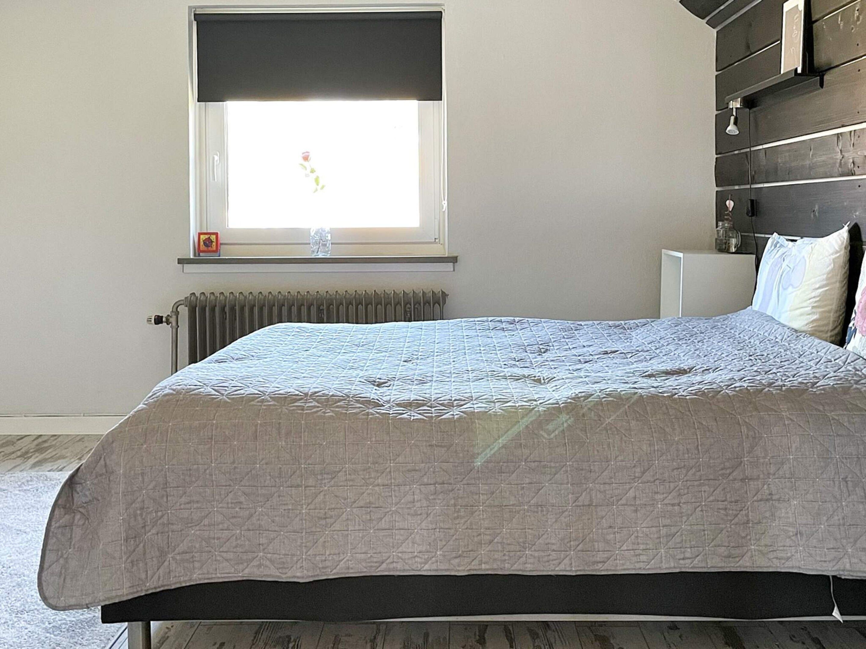 A bedroom with a bed, a radiator, a window with a view, and a shelf with items.
