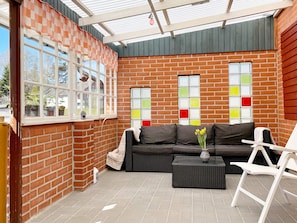 A covered patio area with a brick wall, a black sofa, a white chair, and a tiled floor.