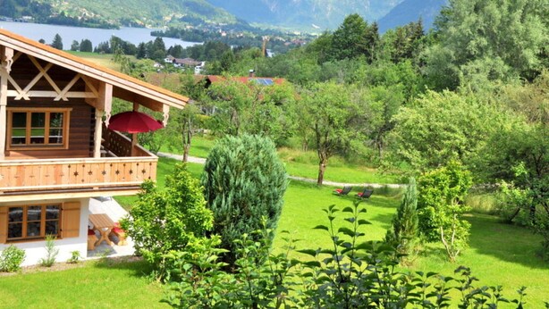 A wooden chalet with a balcony, surrounded by lush greenery and mountains in the background.