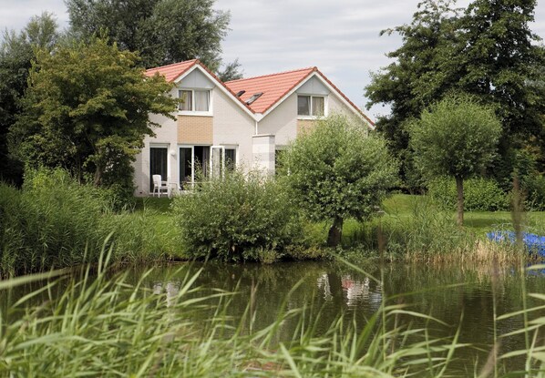 Une maison avec un toit rouge, entourée de verdure et bordée d’un étang.