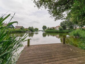 A calm river with a wooden dock in the foreground, surrounded by greenery and houses on the banks. - Nice Holiday Home on the Water in Friesland (Makkum)