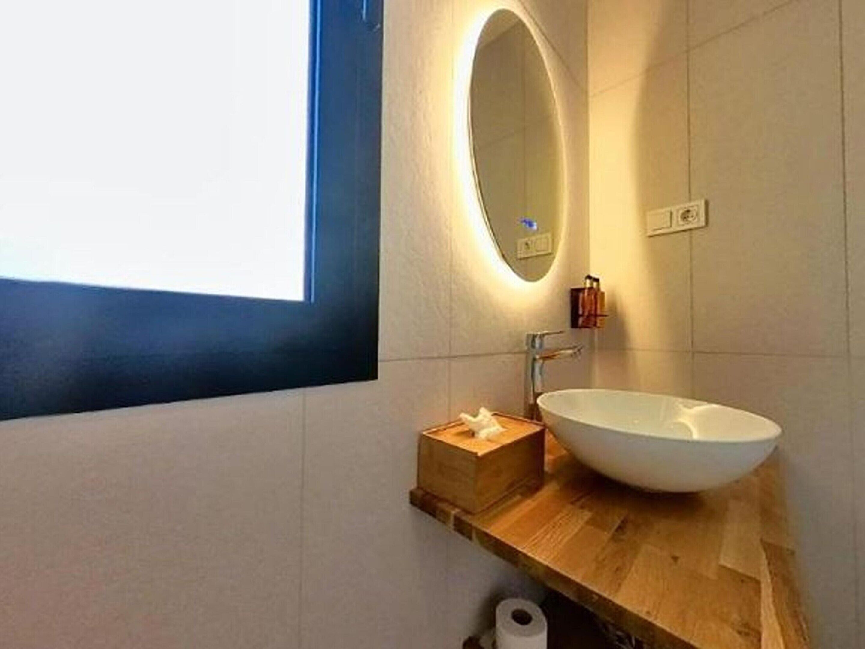 Un baño moderno con un espejo ovalado grande, un lavamanos blanco y un mueble de tocador de madera.