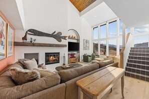 A modern living room with a fireplace, a large window, and a staircase. - Rustic Mountainside 5 Bdrm Ski Home w/ Stunning Views, Private Balconies and Natural Light-Filled Entertainment Space with Access to Grand Hyatt Vail (Vail)