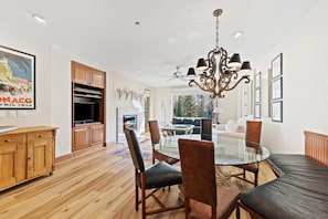 A spacious living area with a glass dining table, comfortable seating, a fireplace, and a large window offering a view of trees.
