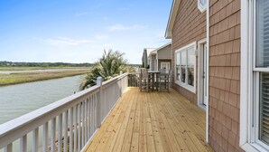 Eine Holzterrasse mit Geländer, die auf einen Fluss blickt, mit Tisch und Stühlen, sowie ein Gebäude mit großen Fenstern.
