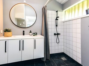 Une salle de bain moderne avec un meuble-lavabo blanc, un miroir rond, une douche équipée d’un pommeau de douche à main et une petite plante en pot posée sur le meuble-lavabo.