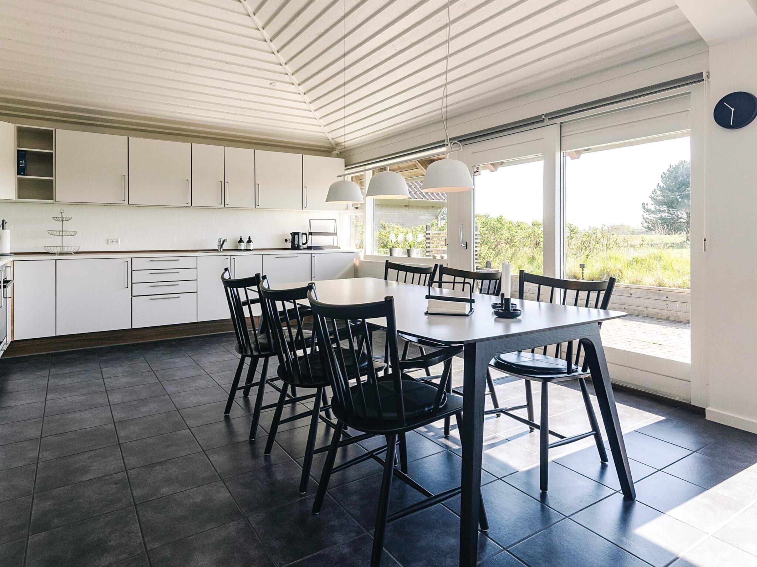 A modern kitchen with white cabinets, a dining table with chairs, and large windows offering a view of greenery.