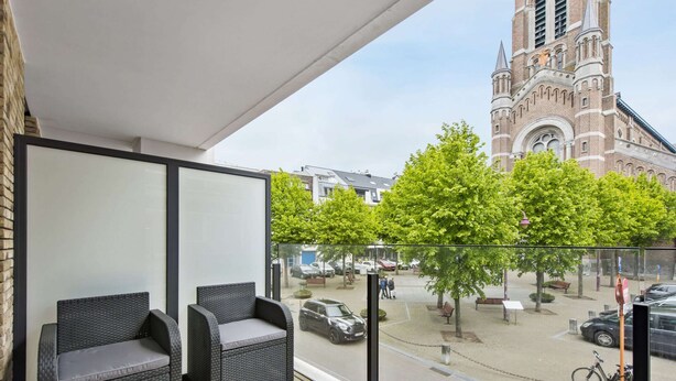 Un balcon agrémenté de meubles en osier, une vue sur une église et une rue bordée d’arbres et traversée par des voitures.