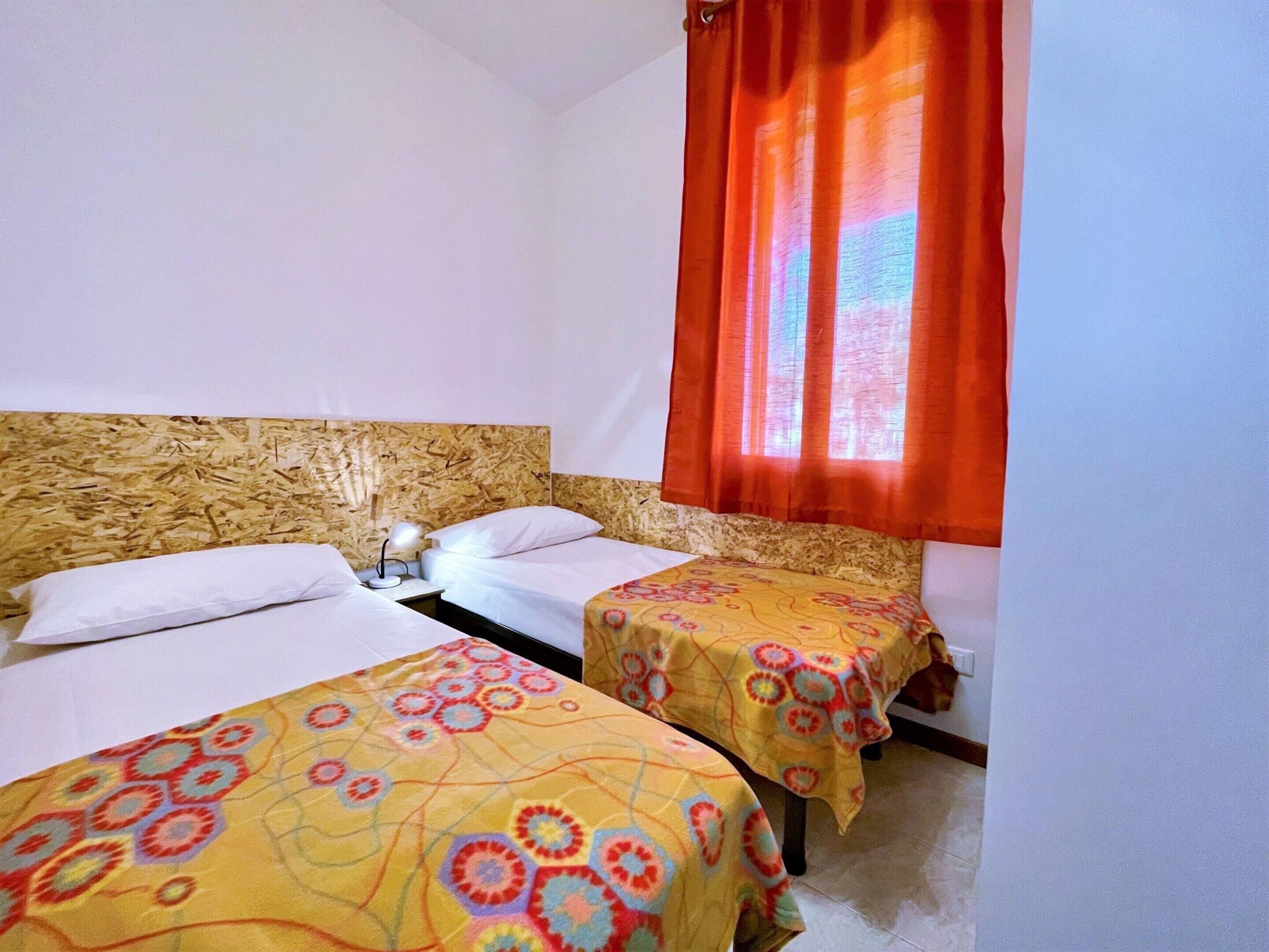 Habitación de hotel con dos camas, cabecero de madera, mesita de noche con lámpara y ventana con cortinas naranjas.