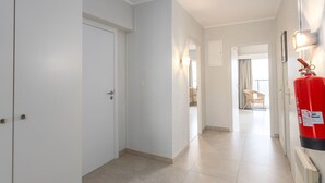 Un couloir avec des portes blanches, un extincteur et une chaise près d’une fenêtre.