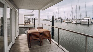 Una terraza techada con mobiliario de madera, una puerta de cristal que da a un edificio blanco y vista a un puerto deportivo con varios barcos.