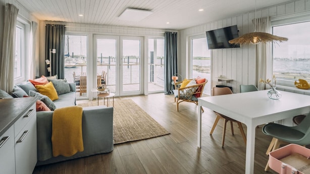 Una sala de estar moderna con sofá, mesa de comedor y sillas, ventanas amplias con vista al agua y un televisor de pantalla plana montado en la pared.