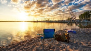 Una playa con una silla y cañas de pescar, un lago tranquilo y una puesta de sol.
