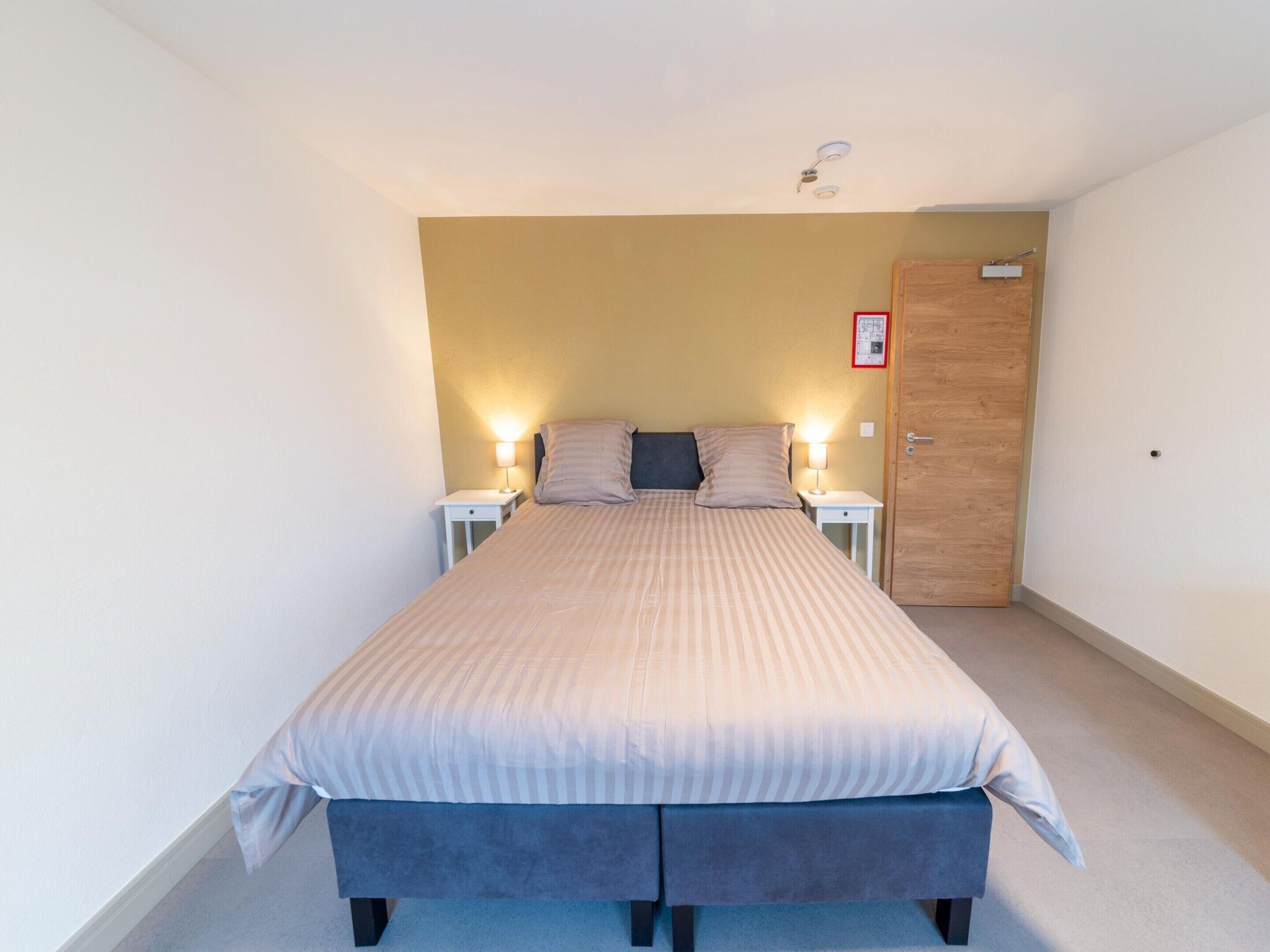 A single bed with a headboard, two bedside tables, a wooden door, and a small wall-mounted light fixture.