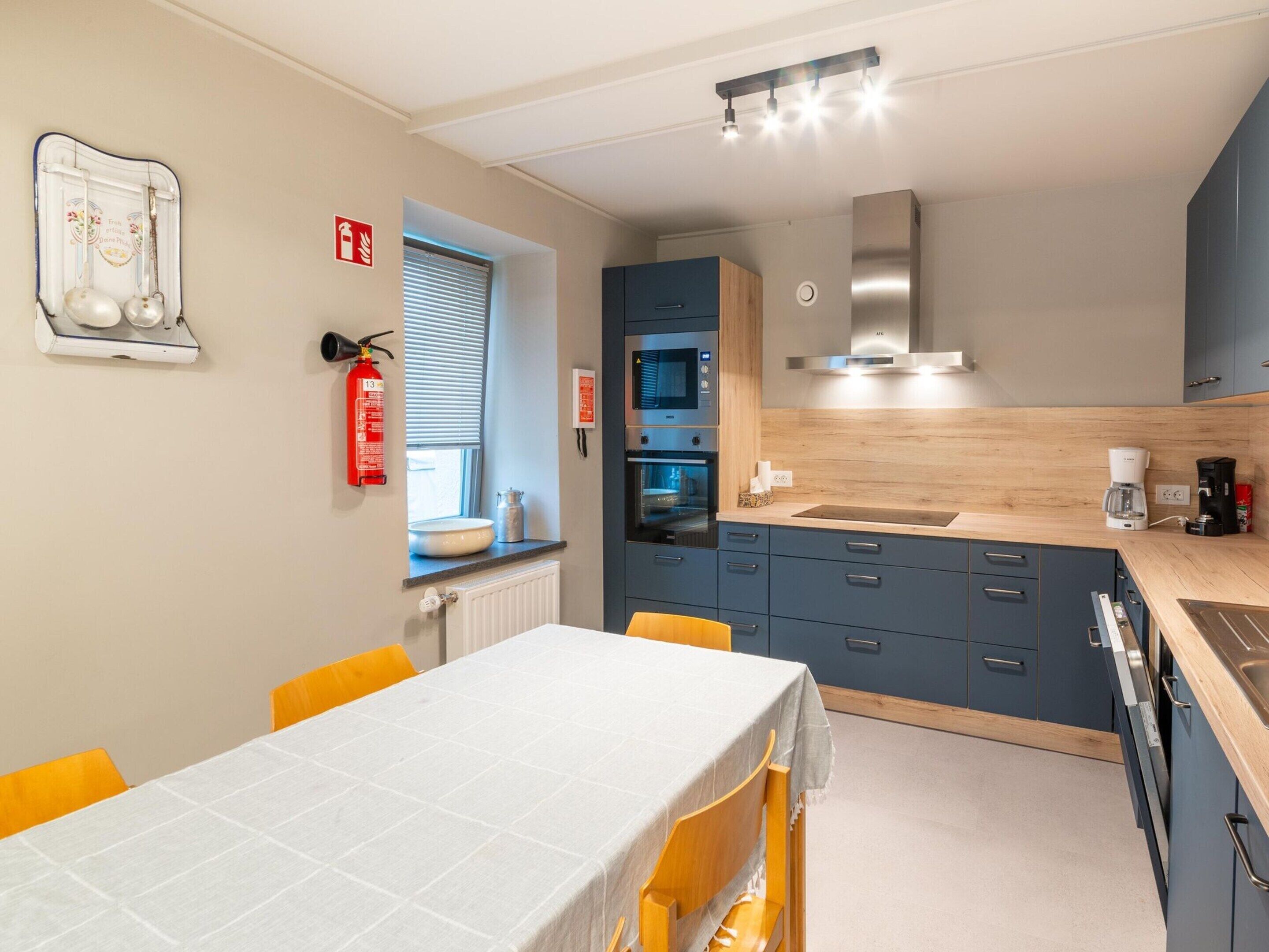 A modern kitchen with a built-in oven, microwave, and coffee maker. There is a dining table with chairs and a fire extinguisher mounted on the wall.