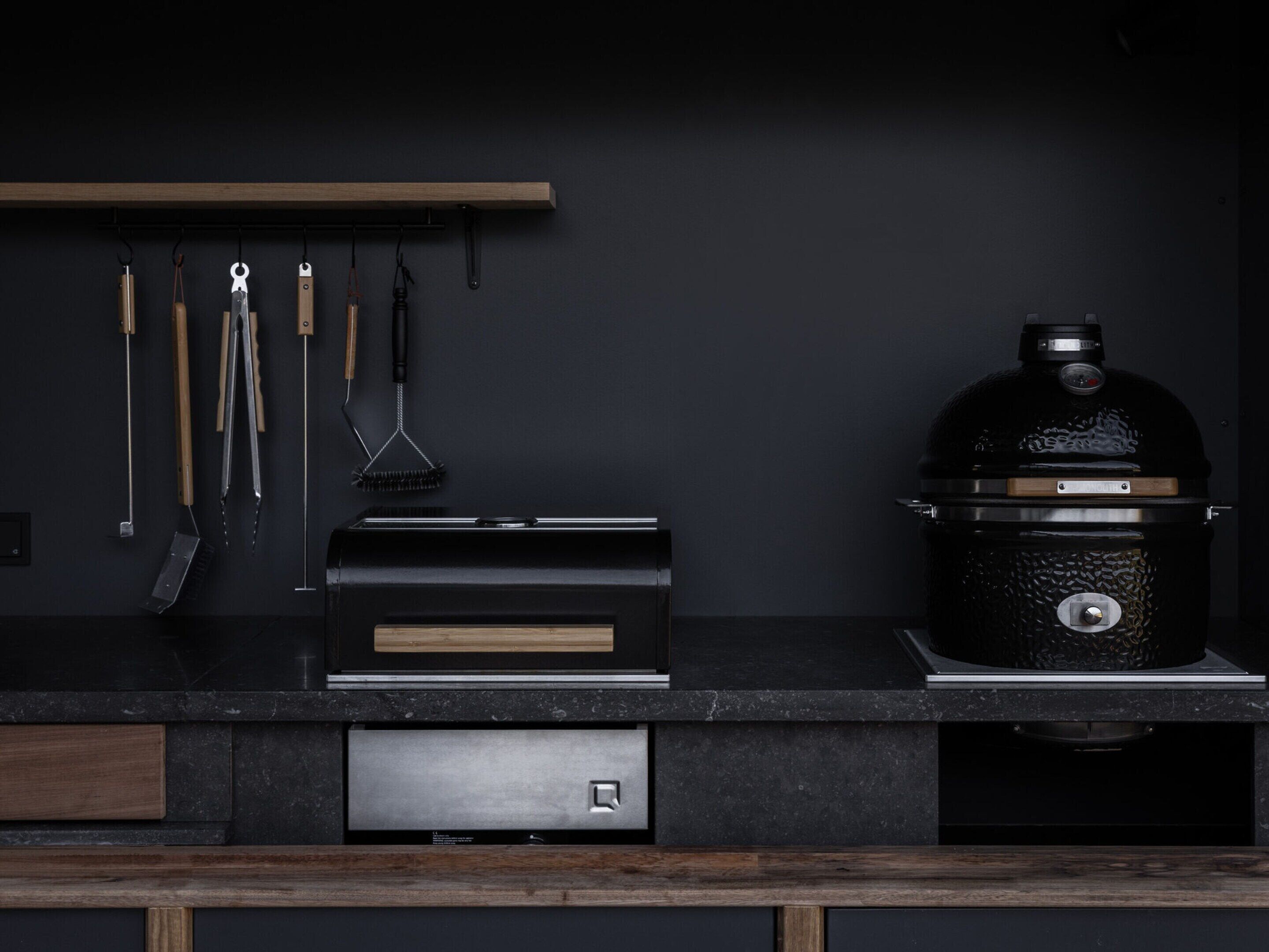 A kitchen with a built-in grill, wooden utensils hanging on the wall, and a drawer unit.