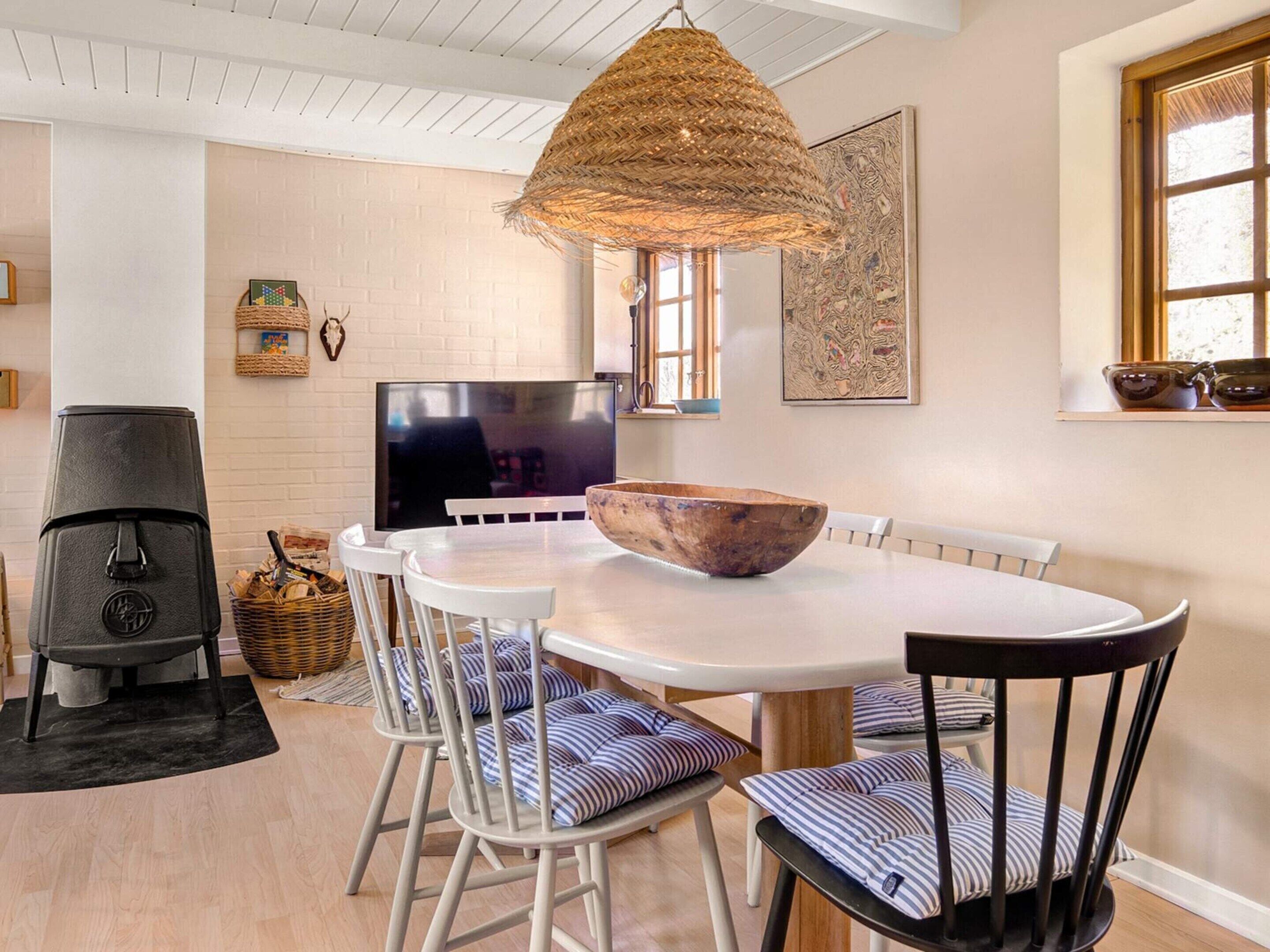 A dining area with a round table, white chairs, a woven hanging light fixture, a television, and a fireplace.