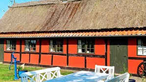 A traditional building with a thatched roof and red walls, featuring a window with a white curtain and a door. - 5 Person Holiday Home in Nexo (Nexø)