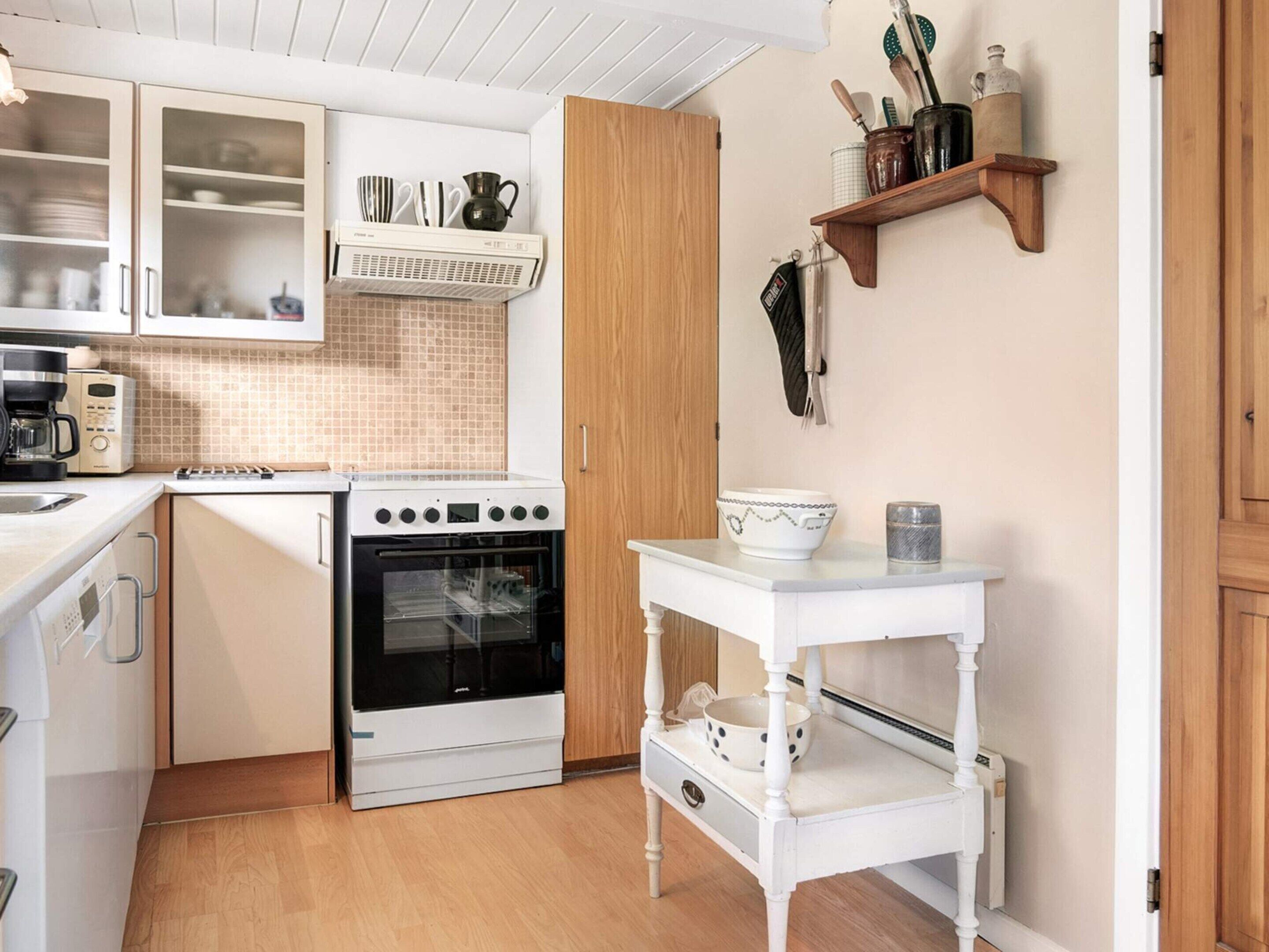 Ett kompakt kök med vita skåp, en inbyggd ugn, en kaffebryggare och ett litet vitt bord med skålar.