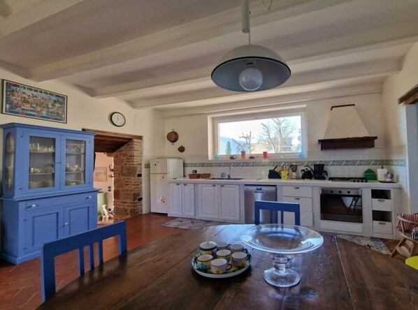 A kitchen with white cabinets, a blue cabinet, a dining table with a clear glass top, and a window with a view of the outdoors. - Villa Lucertole (Città di Castello)