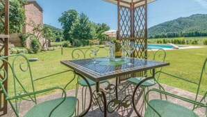 A covered outdoor seating area with a table and chairs, a bottle of wine, and a view of a swimming pool and mountains in the background. - Villa Lucertole (Città di Castello)