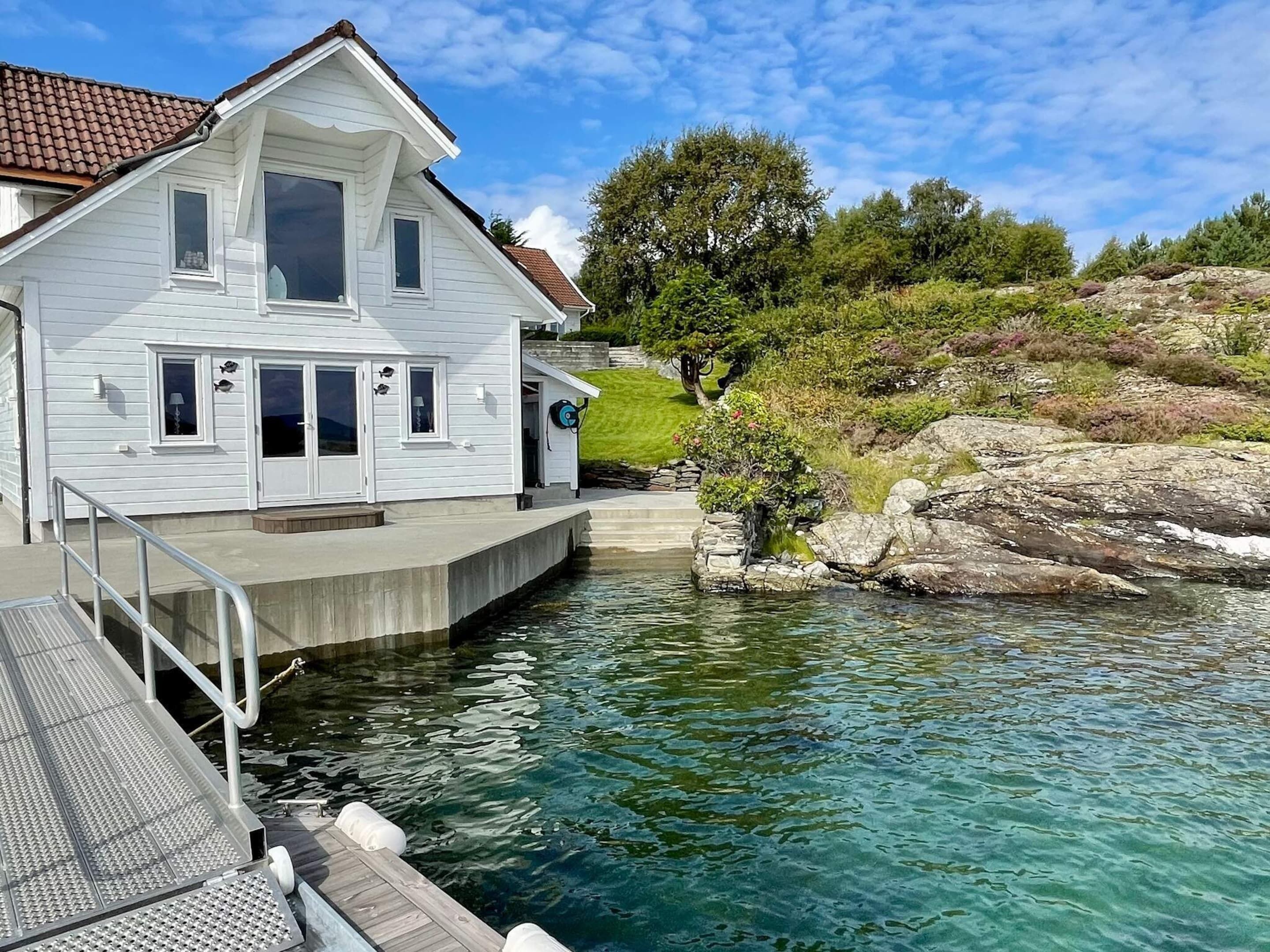 A two-story white house with a balcony, a dock extending into a clear lake, and a rocky hillside in the background.
