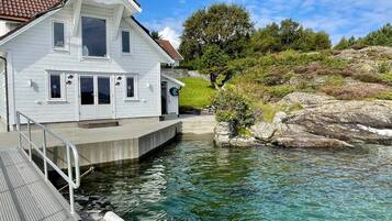 A two-story white house with a balcony, a dock extending into a clear lake, and a rocky hillside in the background.
