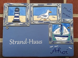 A decorative tile with illustrations of a lighthouse, a seagull, and a sailboat, labeled "Strand-Huus" and "Ahoi". - Holiday Home Strand-huus (Norden)