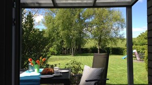 Een terras buiten met een tafel voor twee personen, een stoel en uitzicht op een gazon met bomen en een speeltuin.