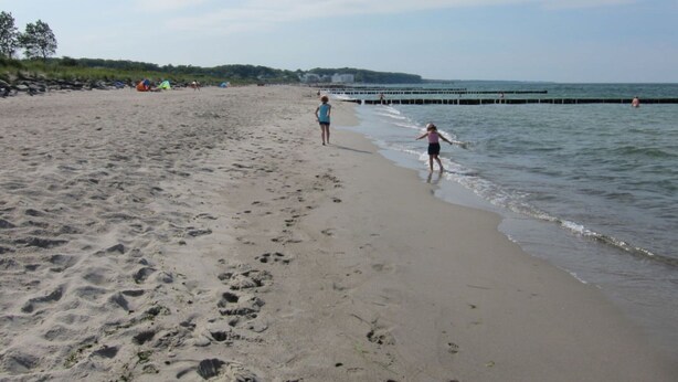 A sandy beach with footprints leading to the water, a pier in the distance, and a clear sky. - Apartment Achtern Meer II (Boergerende-Rethwisch)