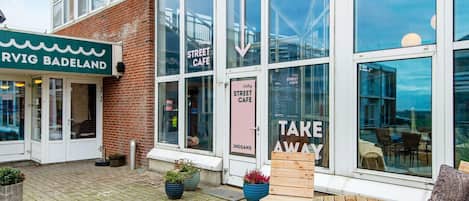 A street cafe with a modern glass facade, outdoor seating with wooden tables and chairs, and a sign indicating 'Take Away' service.