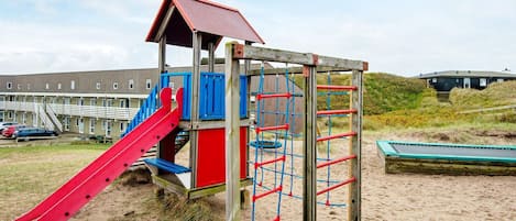 A playground with a slide and climbing structures, set against a backdrop of a building and sandy terrain.