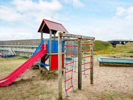 Un parque infantil con un tobogán y estructuras de escalada, con un edificio y terreno arenoso de fondo.