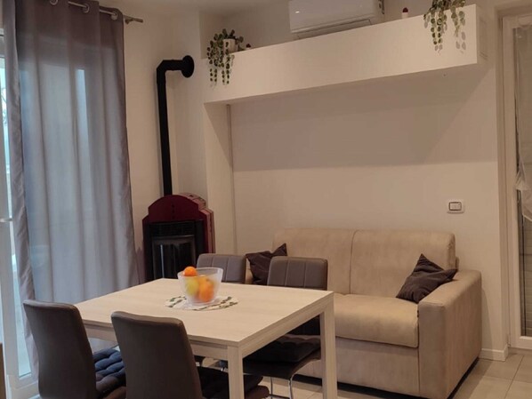 A modern living room with a white sofa, a dining table with chairs, a fireplace, and a ceiling-mounted air conditioning unit.