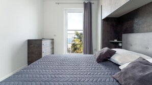 A modern bedroom with a large bed, a dresser, and a view of palm trees through a window. - Apartment Baricevic (Opatija)