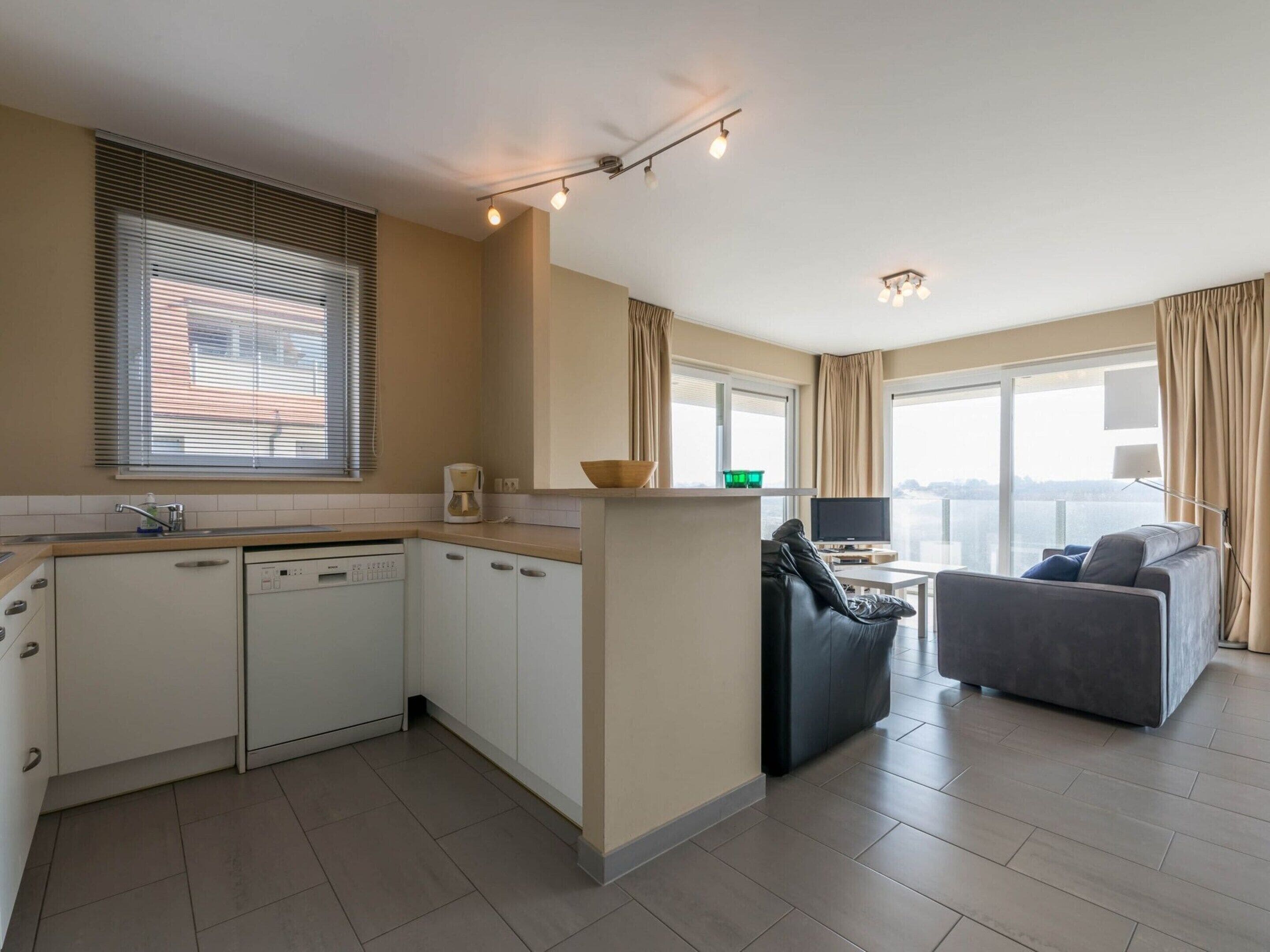 Una cocina moderna con armarios blancos, lavavajillas incorporado y ventana con persianas. Hay un comedor con un sofá de cuero negro y un sofá gris. La habitación tiene un ventanal con cortinas y vistas a la ciudad.