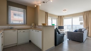 Et moderne køkken med hvide skabe, en indbygget opvaskemaskine og et vindue med persienner. Der er et spiseområde med en sort lædersofa og en grå sofa. Rummet har et stort vindue med gardiner og udsigt til byen.