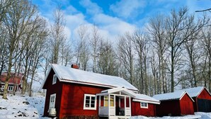 Uma casa de madeira vermelha com telhado coberto de neve, rodeada por árvores e um céu claro.